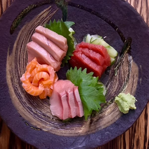 Assorted sashimi, including salmon and tuna, with wasabi and green leaves on a dark plate.