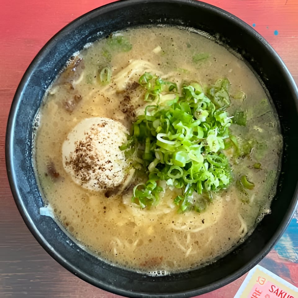 A close-up of a bowl of Bar Bushido ramen with broth, noodles, a soft-boiled egg, and green onions.