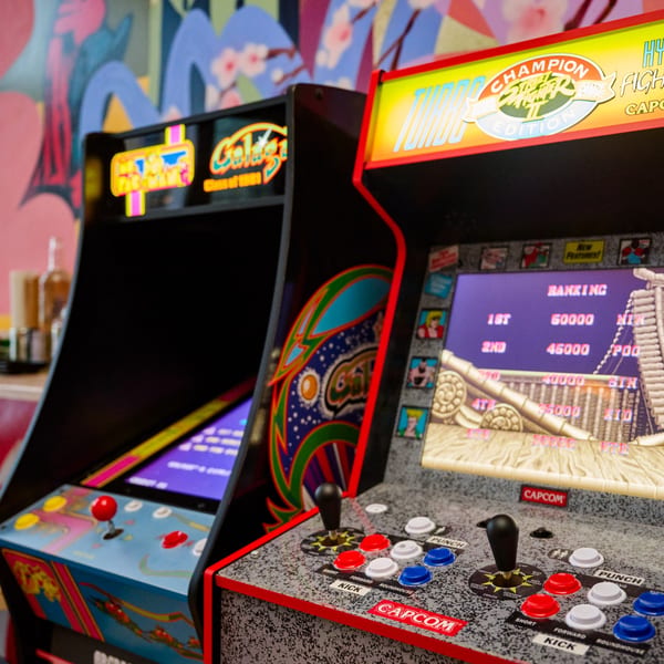 Two vintage arcade machines, one featuring Street Fighter II, against a vibrant, colorful wall mural.