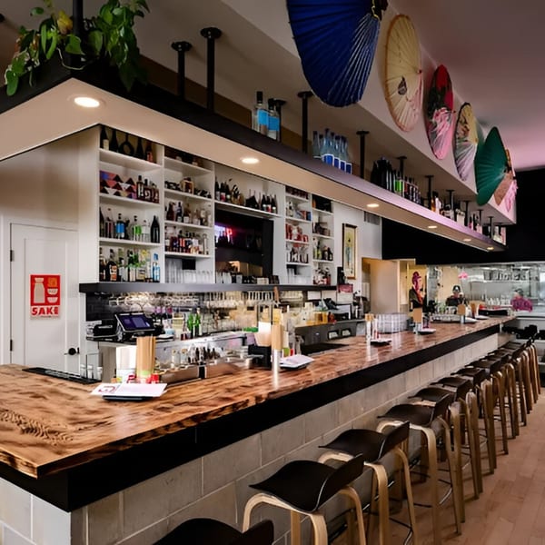 Long wooden bar with black stools, shelves of liquor, and decorative Japanese umbrellas overhead at Bar Bushido.