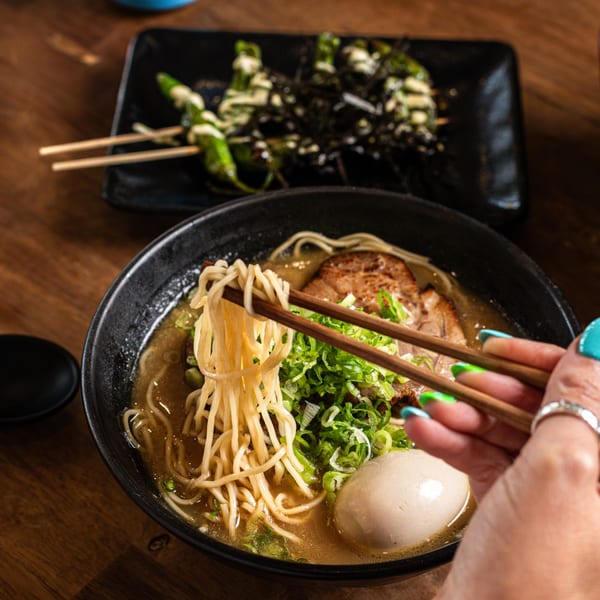 Chopsticks lifting noodles from a bowl of ramen with pork, egg, and scallions, with skewers in the background.