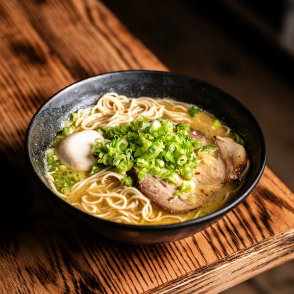 A bowl of Bar Bushido ramen with noodles, pork, egg, and green onions on a wooden table.