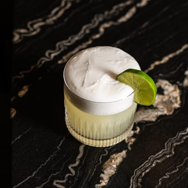A light green cocktail with a thick white foam and a lime wedge on a dark marble surface.