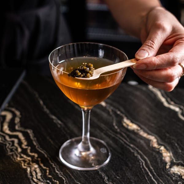 A hand garnishes a Japanese-inspired cocktail with caviar on a wooden spoon over a dark marble bar.