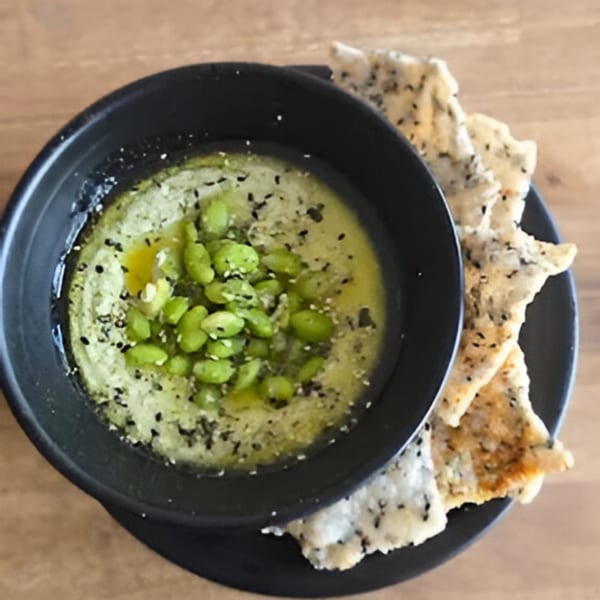 Edamame hummus with olive oil, sesame seeds, and crispy rice crackers on a black plate.
