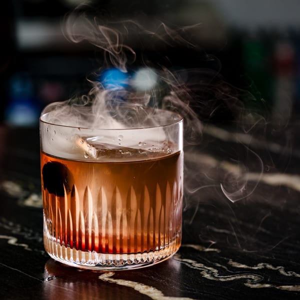 Smoky amber cocktail with a dark cherry and ice cube in a cut glass on a dark marble surface.